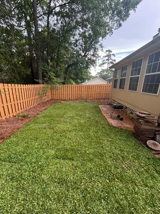 Fenced backyard of a yellow house with freshly laid sod and a mulched border along the wood fence