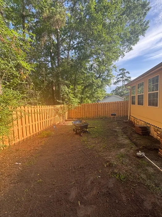 Fenced backyard of a yellow house with bare dirt, sparse weeds and a wheelbarrow before sod installation