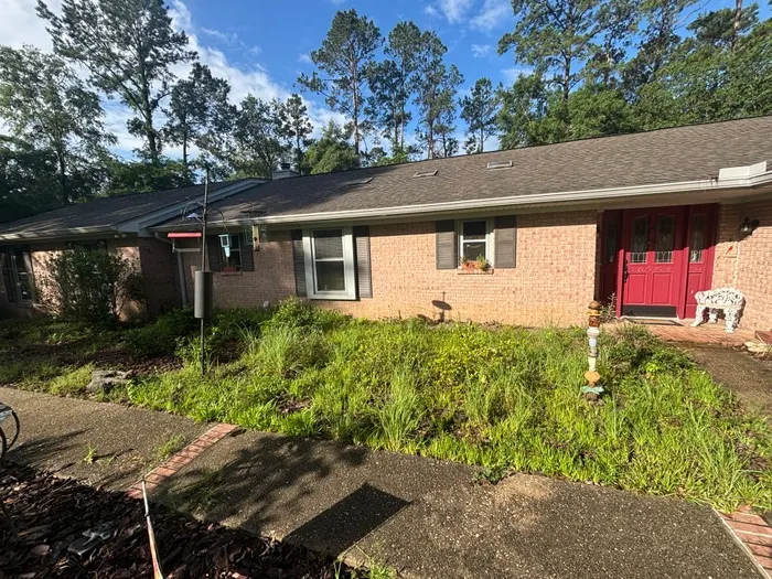 Front of a tan brick ranch with a red door, beds overgrown with tall weeds and grass up to the walkway